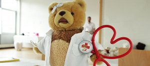Ein rotes, herzförmiges Stethoskop mit einem Button, darauf ein Teddybär mit der Aufschrift „Teddybärkrankenhaus Rosenheim“.