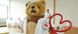 Ein rotes, herzförmiges Stethoskop mit einem Button, darauf ein Teddybär mit der Aufschrift „Teddybärkrankenhaus Rosenheim“.