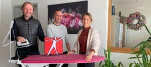 Drei Erwachsene, zwei Männer und eine Frau, stehen in einem hellen Zimmer und halten ein rotes Männchen-Logo; im Hintergrund Fenster und ein großes Blumenbild an der Wand.