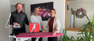 Drei Erwachsene, zwei Männer und eine Frau, stehen in einem hellen Zimmer und halten ein rotes Männchen-Logo; im Hintergrund Fenster und ein großes Blumenbild an der Wand.