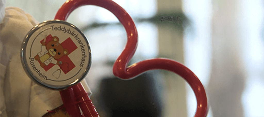 Ein rotes, herzförmiges Stethoskop mit einem Button, darauf ein Teddybär mit der Aufschrift „Teddybärkrankenhaus Rosenheim“.