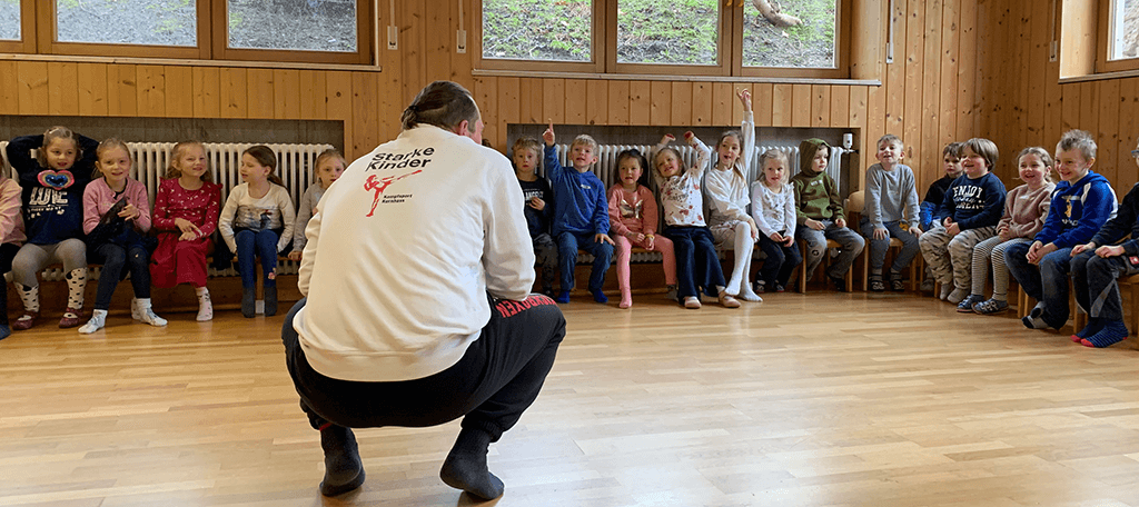Kinder im Hintergrund. Im Vordergrund (mittig) sitzt ein Mann in der Hocke.
