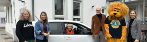 Eine Gruppe von fünf Erwachsenen steht draußen vor einem weißen Auto und einem Gebäude. Unter ihnen ist eine Person im Löwenmaskottchen-Kostüm des Lions Clubs. Eine ältere Frau sitzt lächelnd am Steuer des Autos.