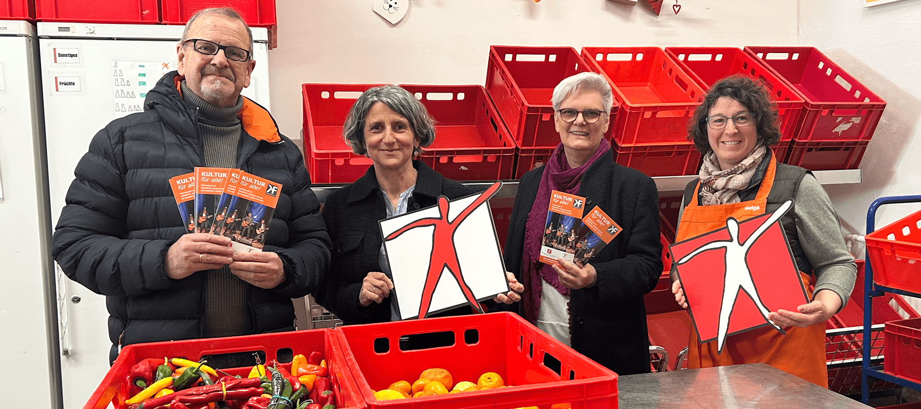 Vier Erwachsene stehen in einem Raum mit roten Kisten voller Gemüse und halten Broschüren sowie Schilder mit einer stilisierten Figur in Rot und Weiß. Alle blicken lächelnd zur Kamera.