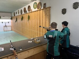 „Zwei Jungen in Schießjacken stehen an einem Schießstand und zielen mit ihren Luftgewehren auf Zielscheiben am anderen Ende des Raums.“