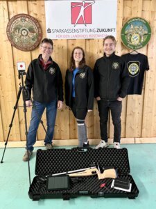 „Drei Mitglieder eines Schützenvereins stehen lächelnd vor einer Holzvertäfelung; vor ihnen liegt ein Luftgewehr in einem offenen Transportkoffer.“