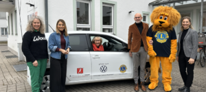 Eine Gruppe von fünf Erwachsenen steht draußen vor einem weißen Auto und einem Gebäude. Unter ihnen ist eine Person im Löwenmaskottchen-Kostüm des Lions Clubs. Eine ältere Frau sitzt lächelnd am Steuer des Autos.