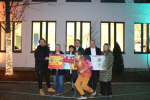 „Sieben Personen stehen vor einem beleuchteten Gebäude, eine Person vorne balanciert auf einem Seil. Zwei Personen halten einen großen bunten Scheck, alle lächeln in die Kamera.“