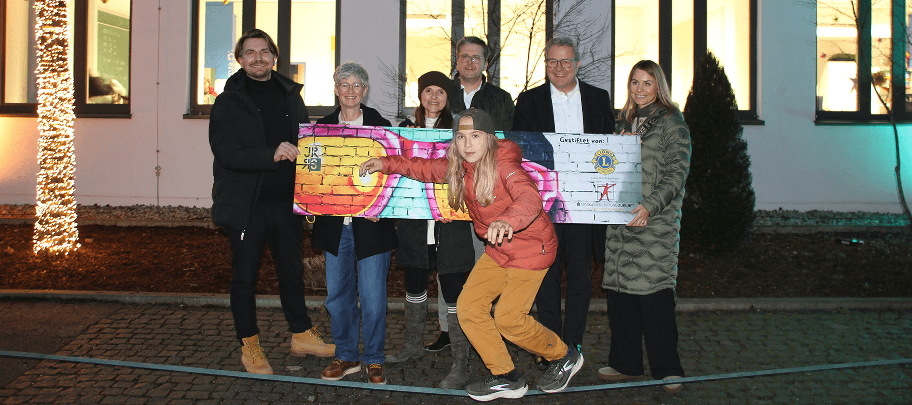 „Sieben Personen stehen vor einem beleuchteten Gebäude, eine Person vorne balanciert auf einem Seil. Zwei Personen halten einen großen bunten Scheck, alle lächeln in die Kamera.“