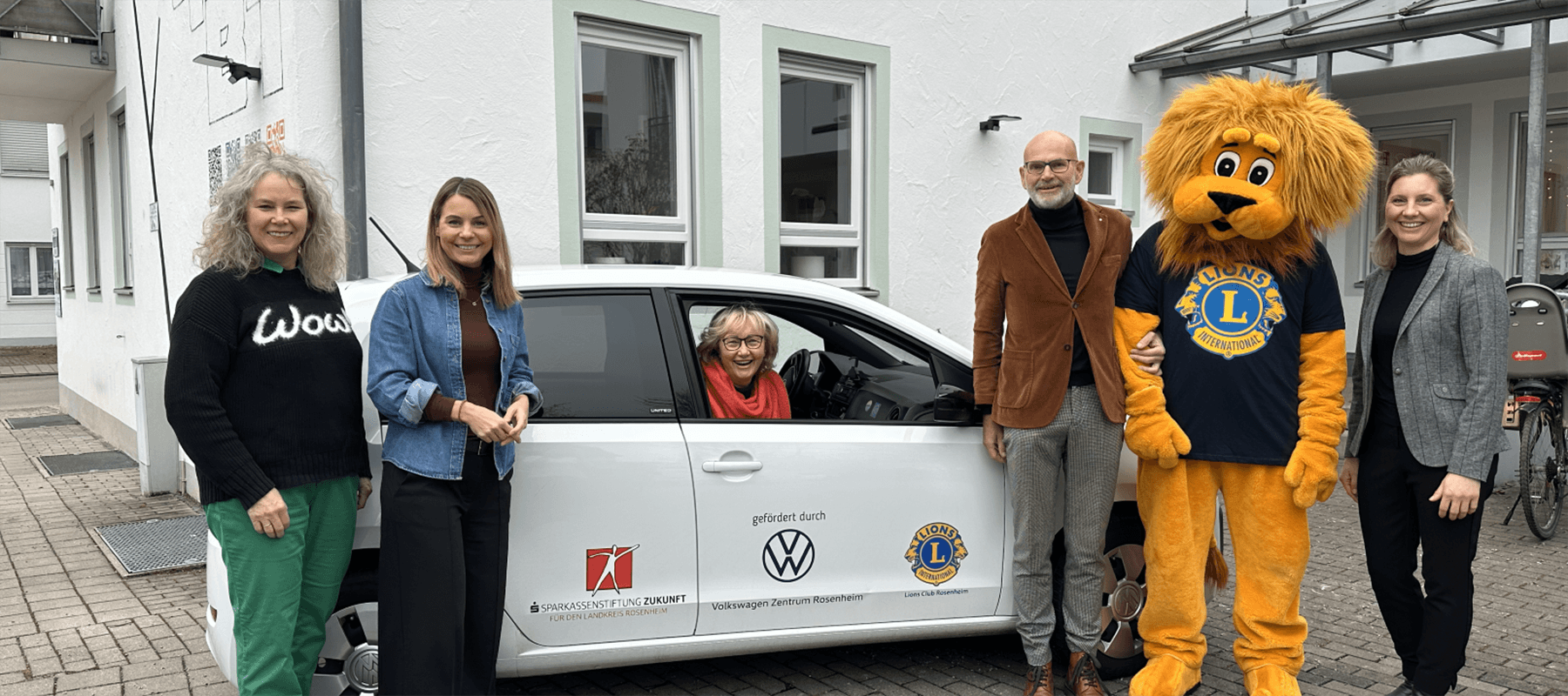 Eine Gruppe von fünf Erwachsenen steht draußen vor einem weißen Auto und einem Gebäude. Unter ihnen ist eine Person im Löwenmaskottchen-Kostüm des Lions Clubs. Eine ältere Frau sitzt lächelnd am Steuer des Autos.