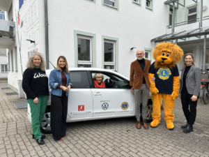 Eine Gruppe von fünf Erwachsenen steht draußen vor einem weißen Auto und einem Gebäude. Unter ihnen ist eine Person im Löwenmaskottchen-Kostüm des Lions Clubs. Eine ältere Frau sitzt lächelnd am Steuer des Autos.