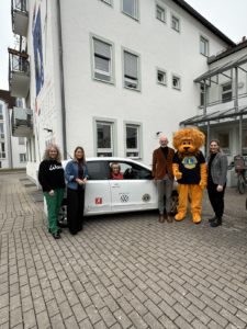 Eine Gruppe von fünf Erwachsenen steht draußen vor einem weißen Auto und einem Gebäude. Unter ihnen ist eine Person im Löwenmaskottchen-Kostüm des Lions Clubs. Eine ältere Frau sitzt lächelnd am Steuer des Autos.